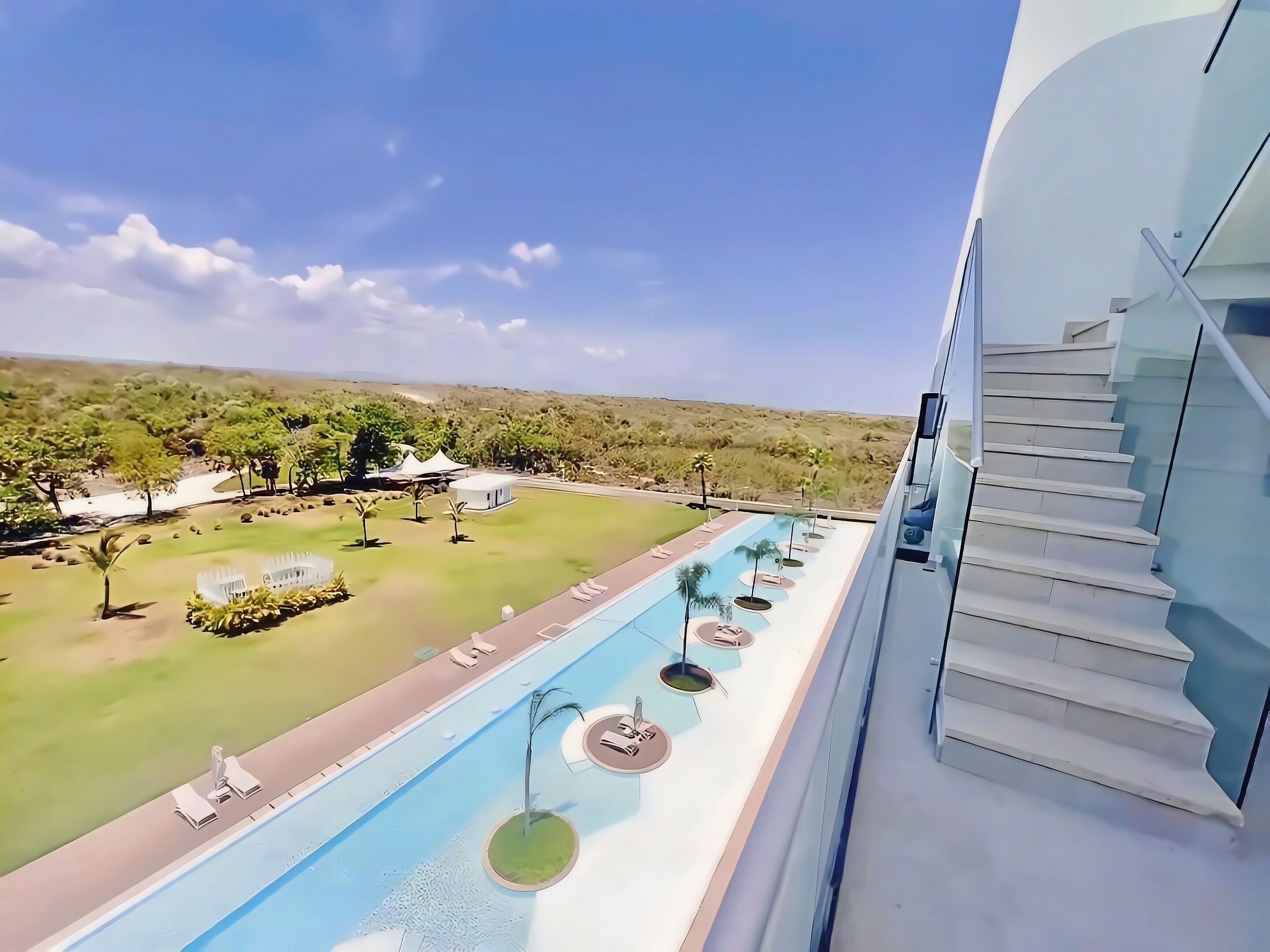 Une piscine avec des chaises longues, un escalier et une vue sur un espace paysager agrémenté d’arbres et d’un bâtiment blanc.