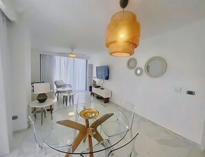 Een moderne hotelkamer met een glazen eettafel, een plafondventilator, een groot raam met gordijnen, een flatscreen televisie en een spiegel aan de muur.