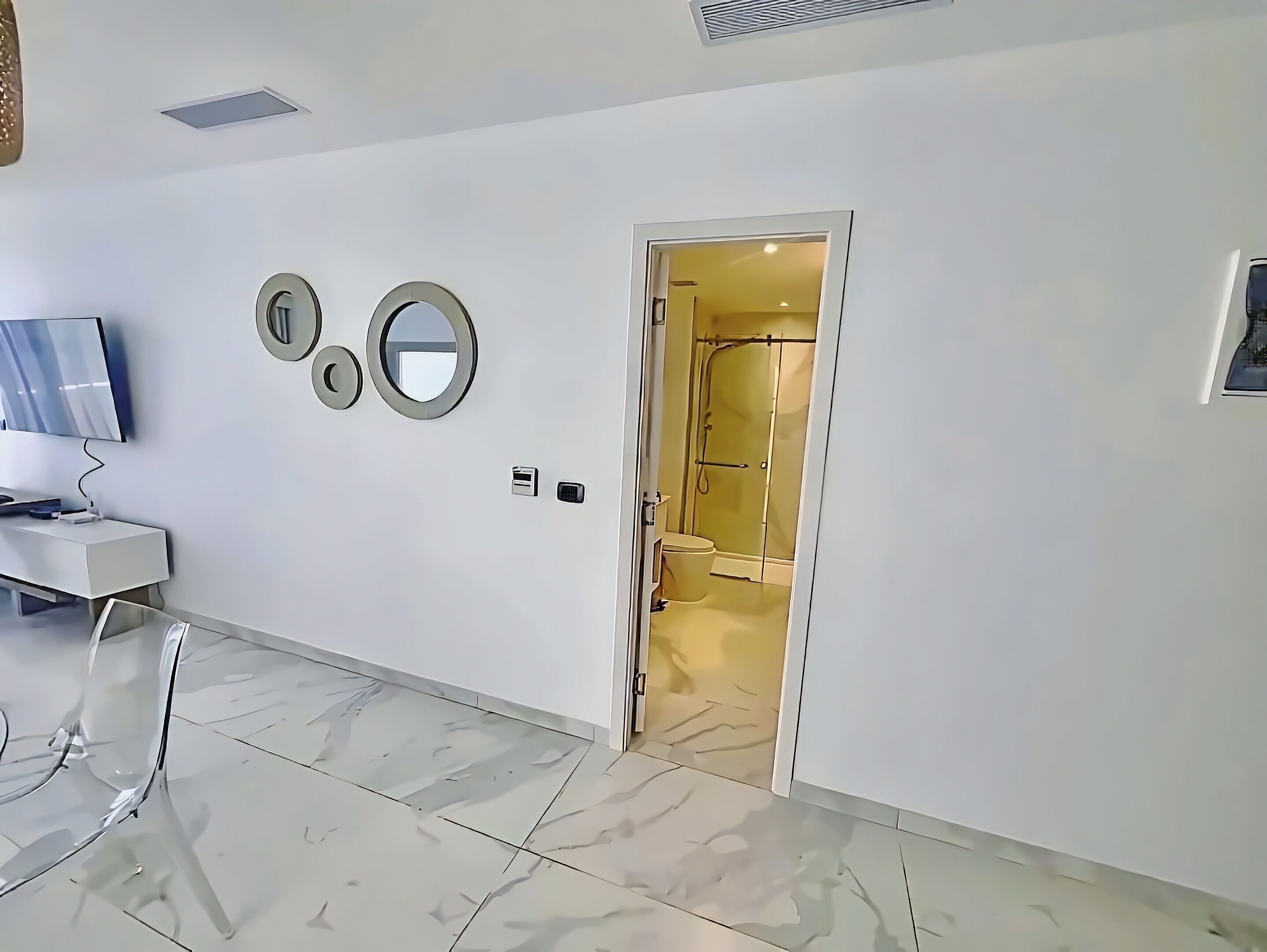 A hotel room with a TV, three wall-mounted mirrors, a door leading to a bathroom, and a tiled floor.