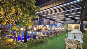 Ein Poolbereich im Freien mit Kunstgrasanlage, umgeben von Tischen und Stühlen, unter einer überdachten Terrasse mit Lichterleiste.