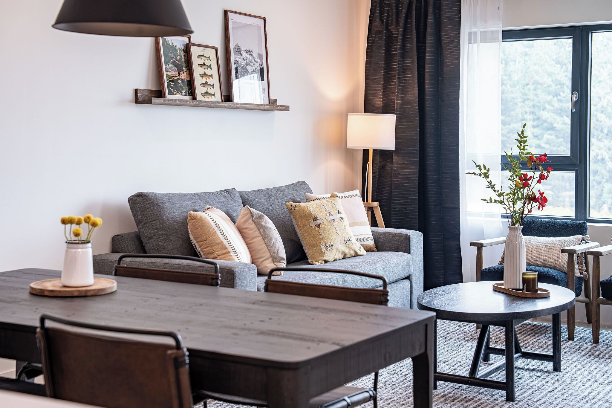 A modern living room with a grey sofa, a round coffee table, and a dining table with chairs. There is a lamp, a vase with flowers, and a window with a view of trees.