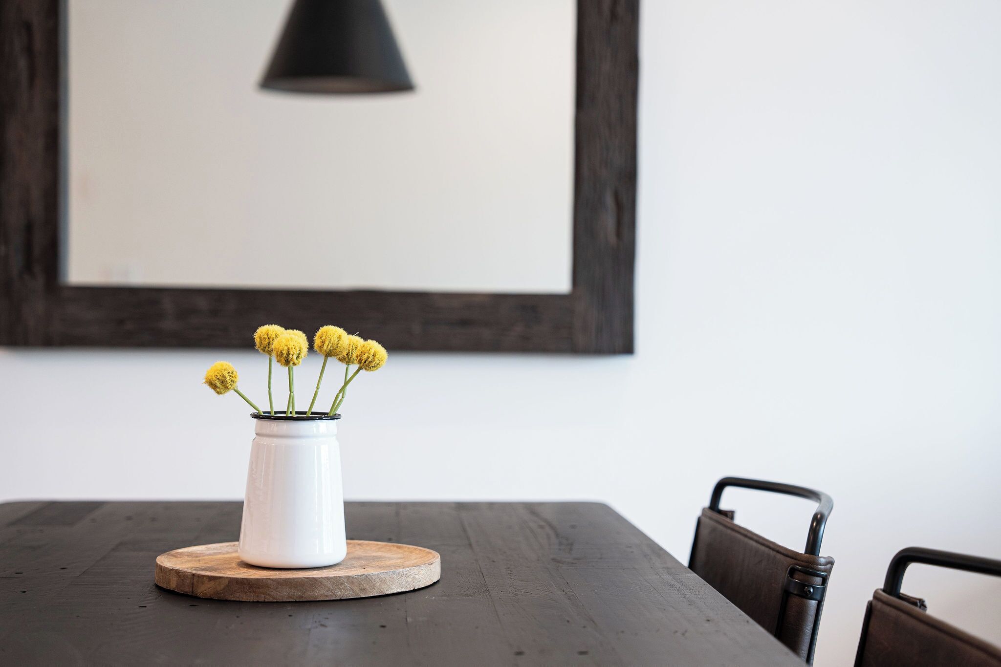 A white vase with yellow flowers on a round wooden coaster on a dark wooden table.