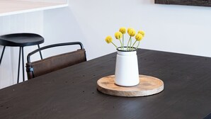 Ein Esstisch mit einer weißen Vase voller gelber Blumen, einer schwarzen Lampe und einem Holzuntersetzer.