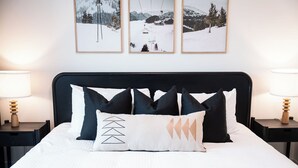 Ein ordentlich bezogenes Bett mit schwarzem Kopfteil, weißer Bettwäsche und dekorativen Kissen. Darüber an der Wand hängen drei gerahmte Bilder.