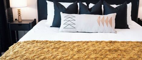 Un lit bien fait, recouvert d’une couverture à motifs, une lampe de chevet et trois tableaux encadrés accrochés au mur.