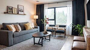 Ein modernes Wohnzimmer mit grauem Sofa, einem kleinen runden Tisch und einem Couchtisch. An der Wand befinden sich gerahmte Bilder, ein Topf mit Pflanze und ein Fenster mit Vorhängen.