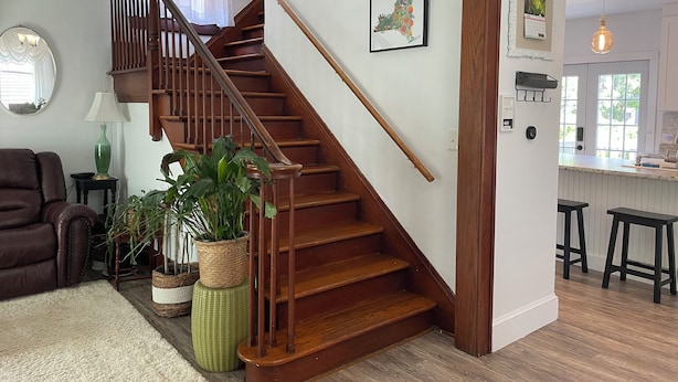Eine Treppe mit hölzernen Geländern führt zu einer oberen Etage. Auf einem Hocker steht eine grüne Pflanze, ein brauner Ledersessel und eine weiße Wand mit einem gerahmten Bild.