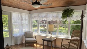 Ein Wintergarten mit Holzboden, großen Fenstern mit Spitzendeckchen, einem Deckenventilator, einem Holzschaukelstuhl, einem Tisch mit Lampe und einer hängenden Pflanze.