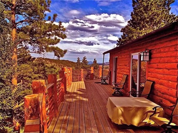 Eine Holzterrasse mit Tisch und Stühlen, eine rote Holzhütte und Blick auf Bäume und Berge.