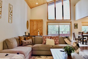 A cozy living room with a sectional sofa, a dining table, and a large window offering a view of trees. - Modern Tahoe cabin: A/C, ping pong, EV charge, family friendly, work trips (Truckee)