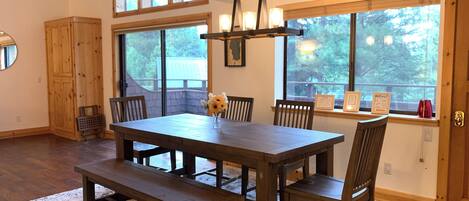Un comedor con mesa de madera y bancos, ventanas amplias y vista a árboles.