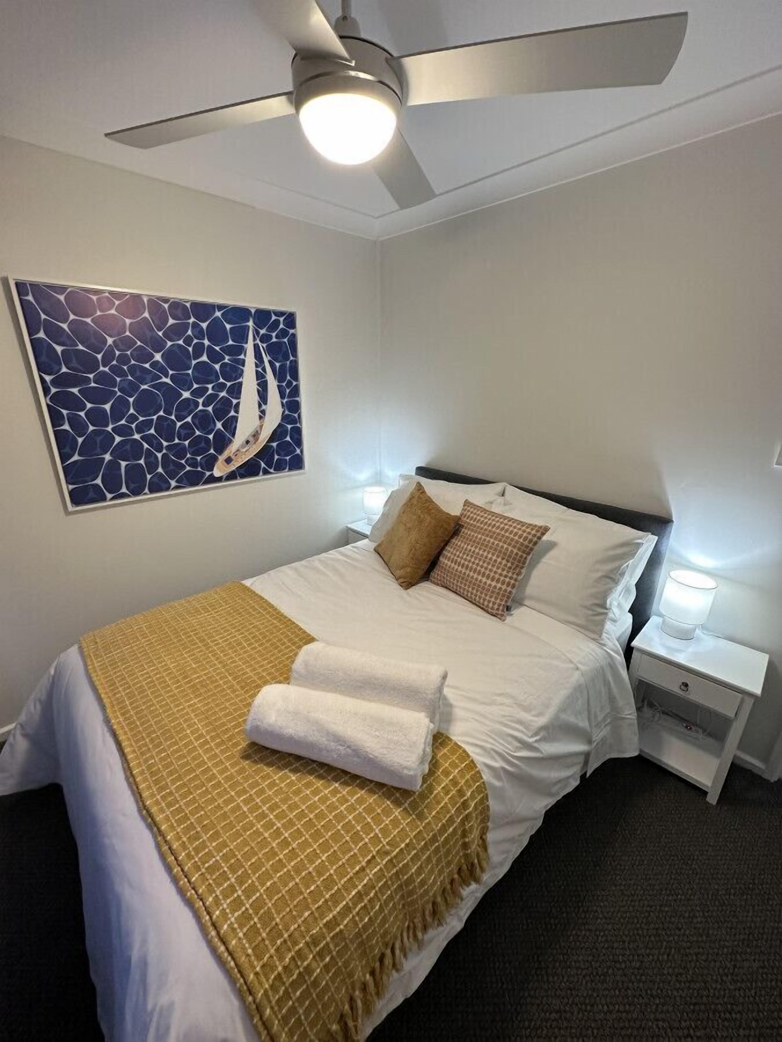 A neatly made bed with a white and gold checkered blanket, a ceiling fan, a wall-mounted artwork, and bedside lamps.