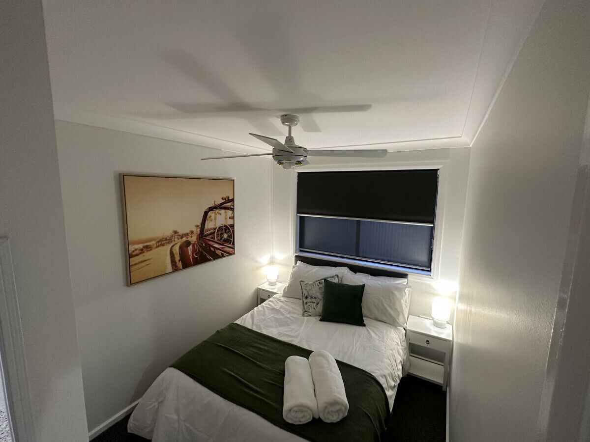 A hotel room with a bed, a ceiling fan, a nightstand with a lamp, and a window with blinds.