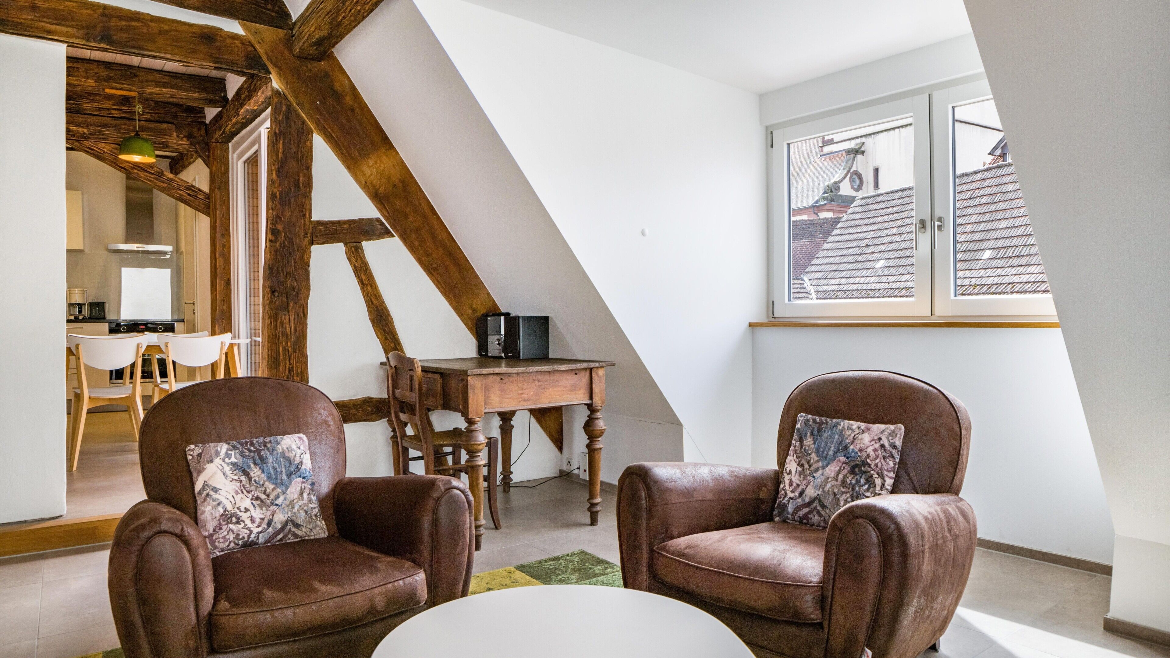 Une pièce de style « loft », avec des poutres en bois apparentes, un coin repas et une grande fenêtre donnant sur un immeuble.