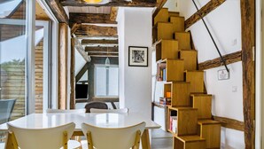 Un intérieur moderne avec un escalier en bois, un coin repas doté d’une table et de chaises blanches, et une bibliothèque.