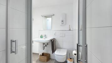A modern bathroom with a glass shower enclosure, a white sink, a toilet, and a small trash bin.