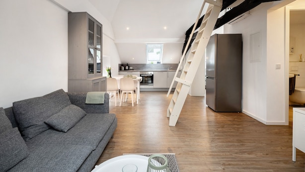 Un apartament modern amb un sofà gris, una zona de menjador, una cuina i una escala.