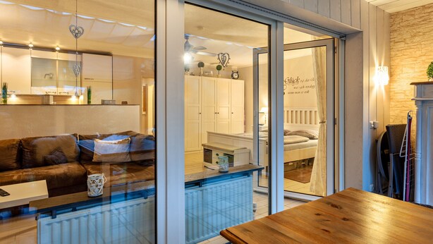 Un interior moderno con un separador de vidrio, una mesa de madera, un sofá y una zona de cocina visible al otro lado.