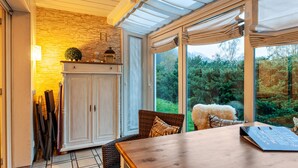Una habitación con una mesa de madera, sillas de mimbre, un armario y vistas a la vegetación a través de amplias ventanas.