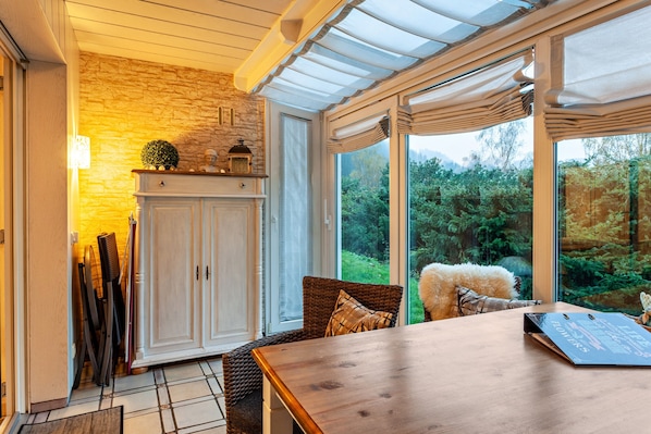 Une pièce avec une table en bois, des chaises en osier, un buffet et une vue sur la verdure grâce à de grandes fenêtres.