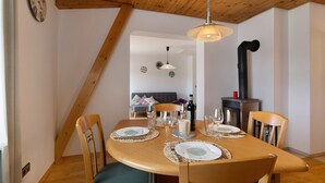 Zona pranzo con tavolo in legno apparecchiato per quattro persone, sedie, lampadario e camino sullo sfondo.