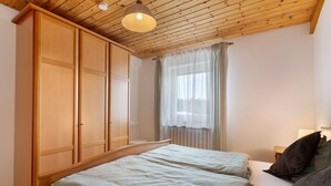 Una camera da letto con un'armadiatura in legno, un letto con lenzuola bianche, una finestra con tende trasparenti e un soffitto in legno.