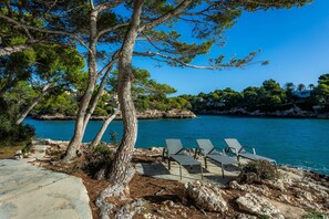 A coastal area with a clear view of the sea, a rocky shoreline, and a few lounge chairs.