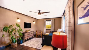A room with a ceiling fan, a flat-screen TV, a bed with a red lamp, a wicker chair, a wooden dresser, and a potted plant. - Renovated Coastal Retreat • Walk to Town • Near Stone Harbor & Other Beaches (Cape May Court House)