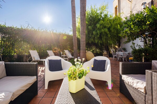 Un patio con mobiliario de mimbre, una mesa central con una planta y un fondo iluminado por el sol.