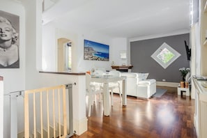 Una sala de estar moderna con comedor, un sofá blanco, una mesa de vidrio y un cuadro de una escena costera.
