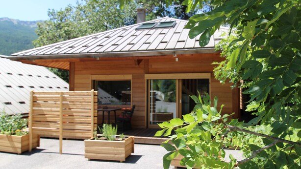 Una cabaña de madera con techo inclinado, un patio con mesa y sillas, y un jardín con jardineras de madera.