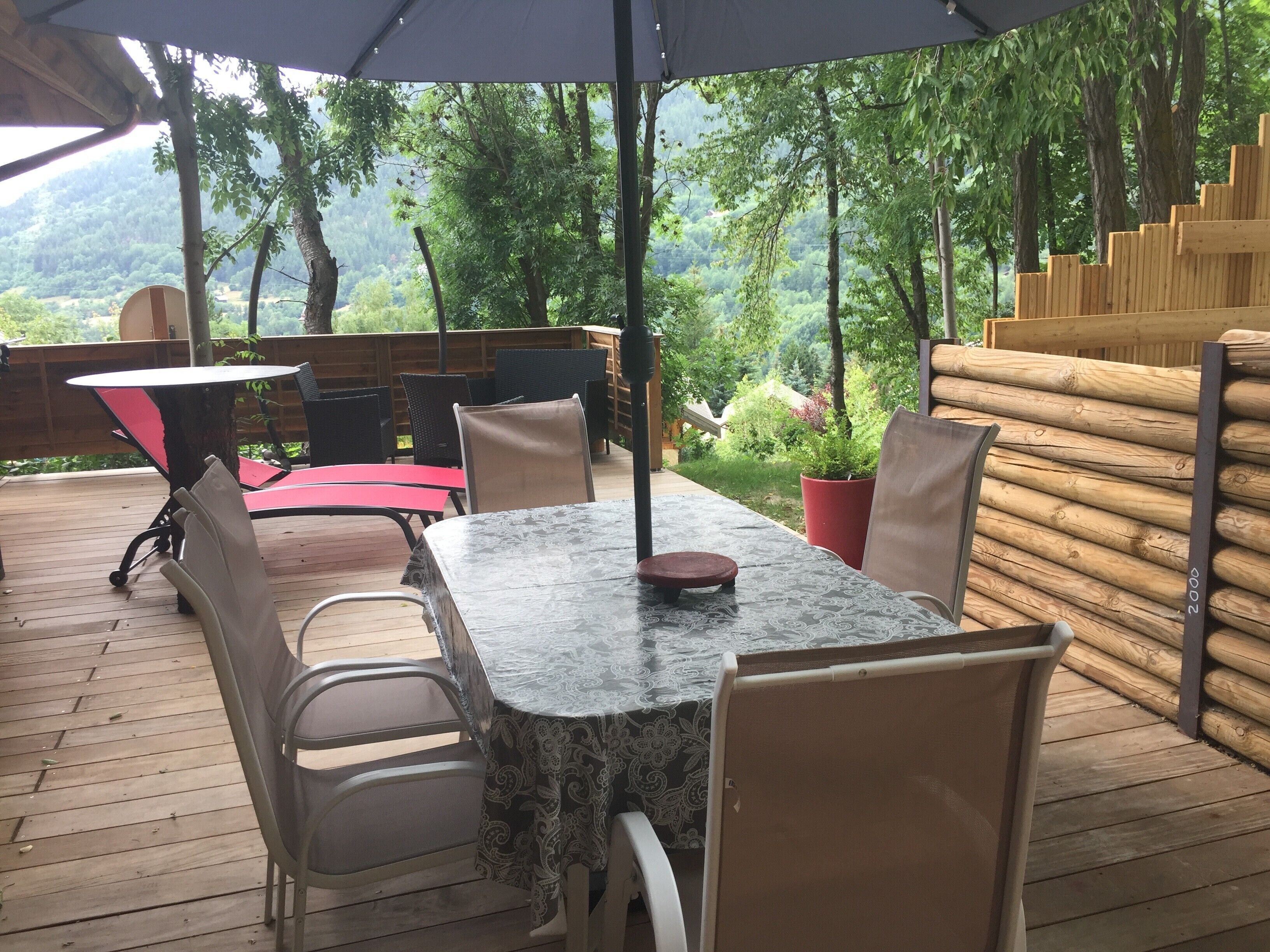 Un patio exterior con mesa y sillas, rodeado por una terraza de madera y vistas a una zona boscosa.