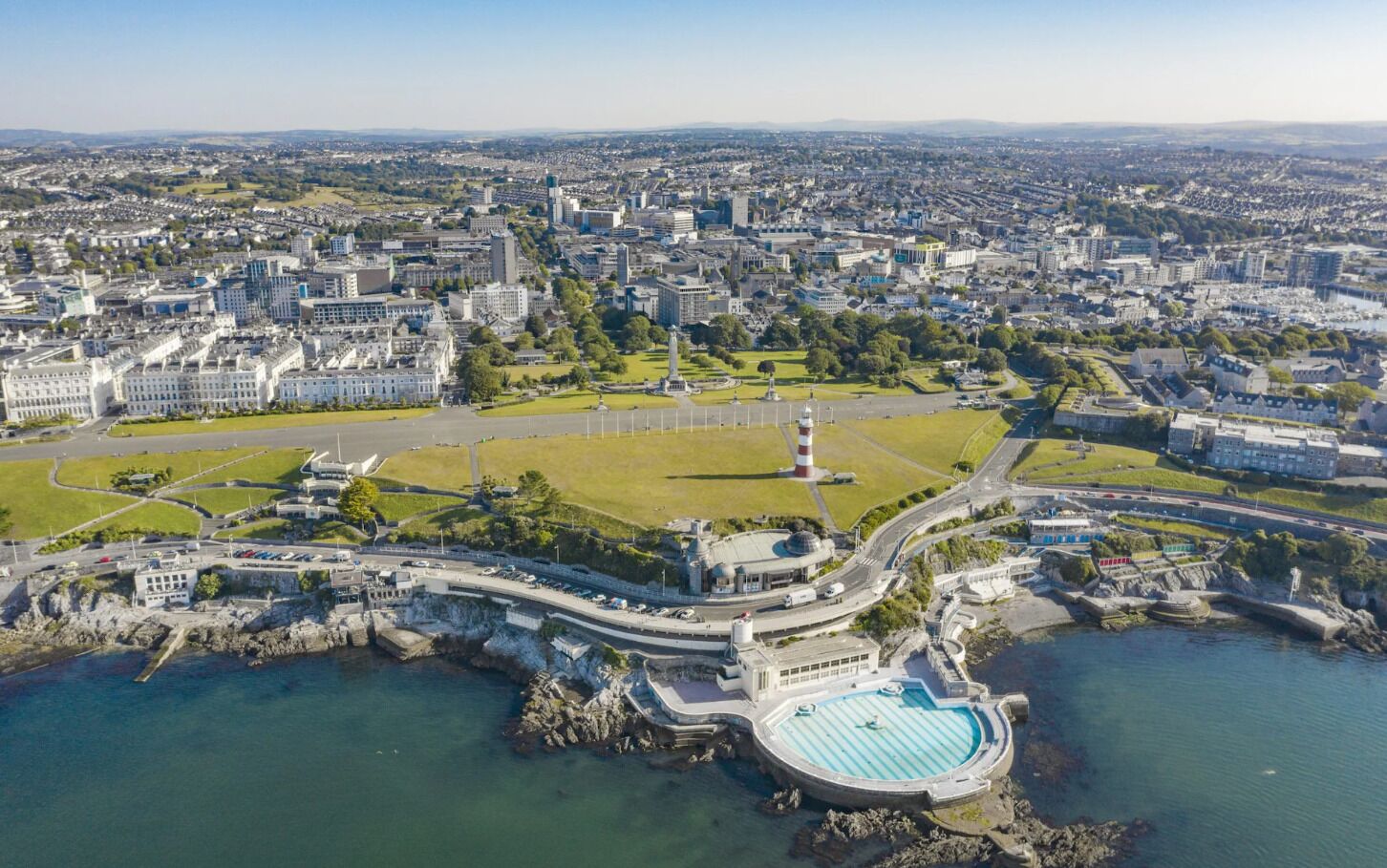 An aerial view of a coastal city with a prominent waterfront promenade, a lighthouse, and a large hotel complex.