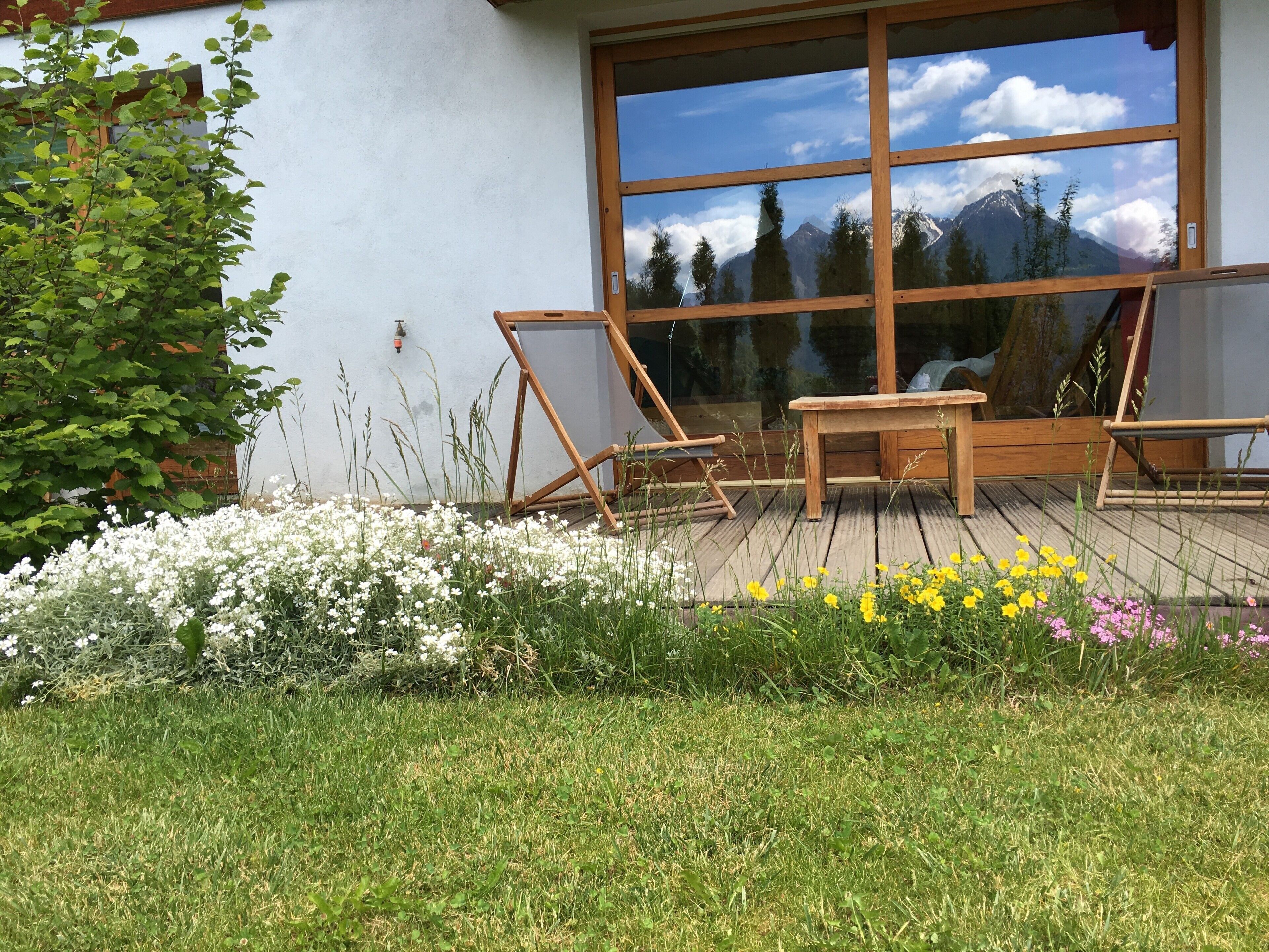 A wooden deck with a table and chairs, a large window reflecting mountains, and a garden with various flowers.