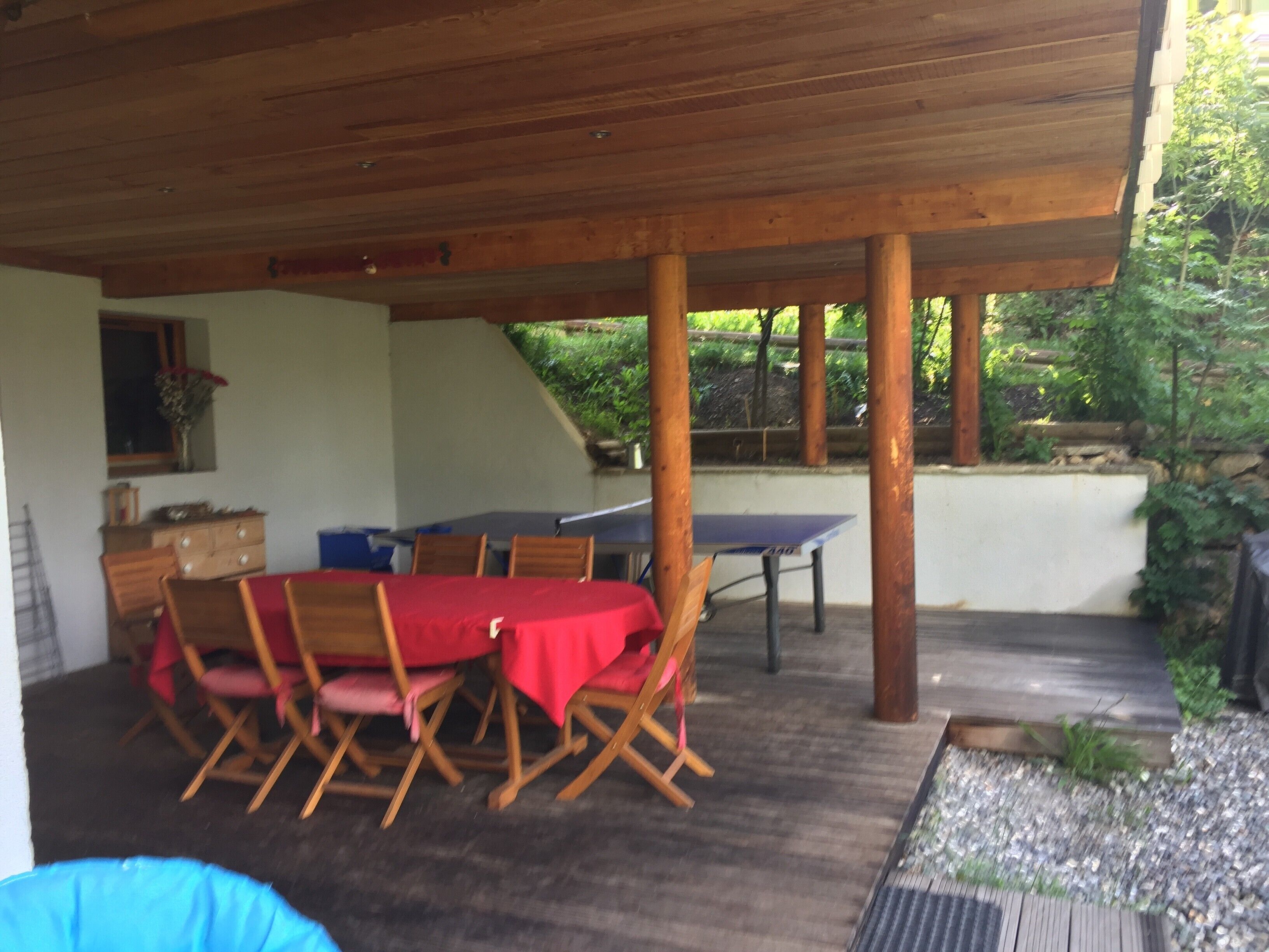 Un patio cubierto al aire libre con mesa de ping pong, sillas de madera y una mesa de comedor con mantel rojo.