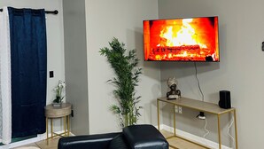 En stue med et fladskærms-tv, en loftsvifte, et sofabord med en plante, en sort lædersofa og et konsolbord med gylden ramme.