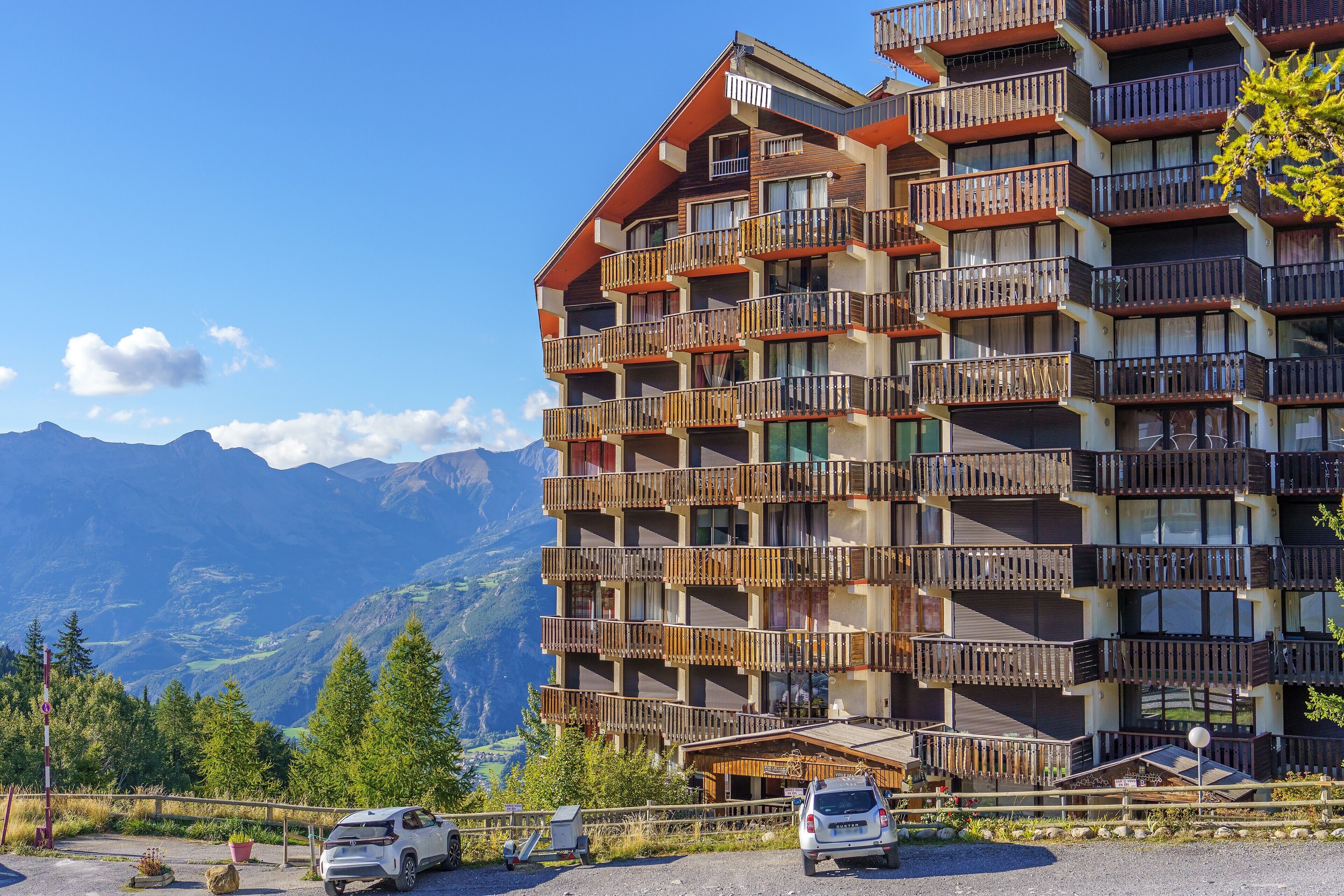 A multi-story hotel with balconies, a parking area, and a mountainous backdrop.