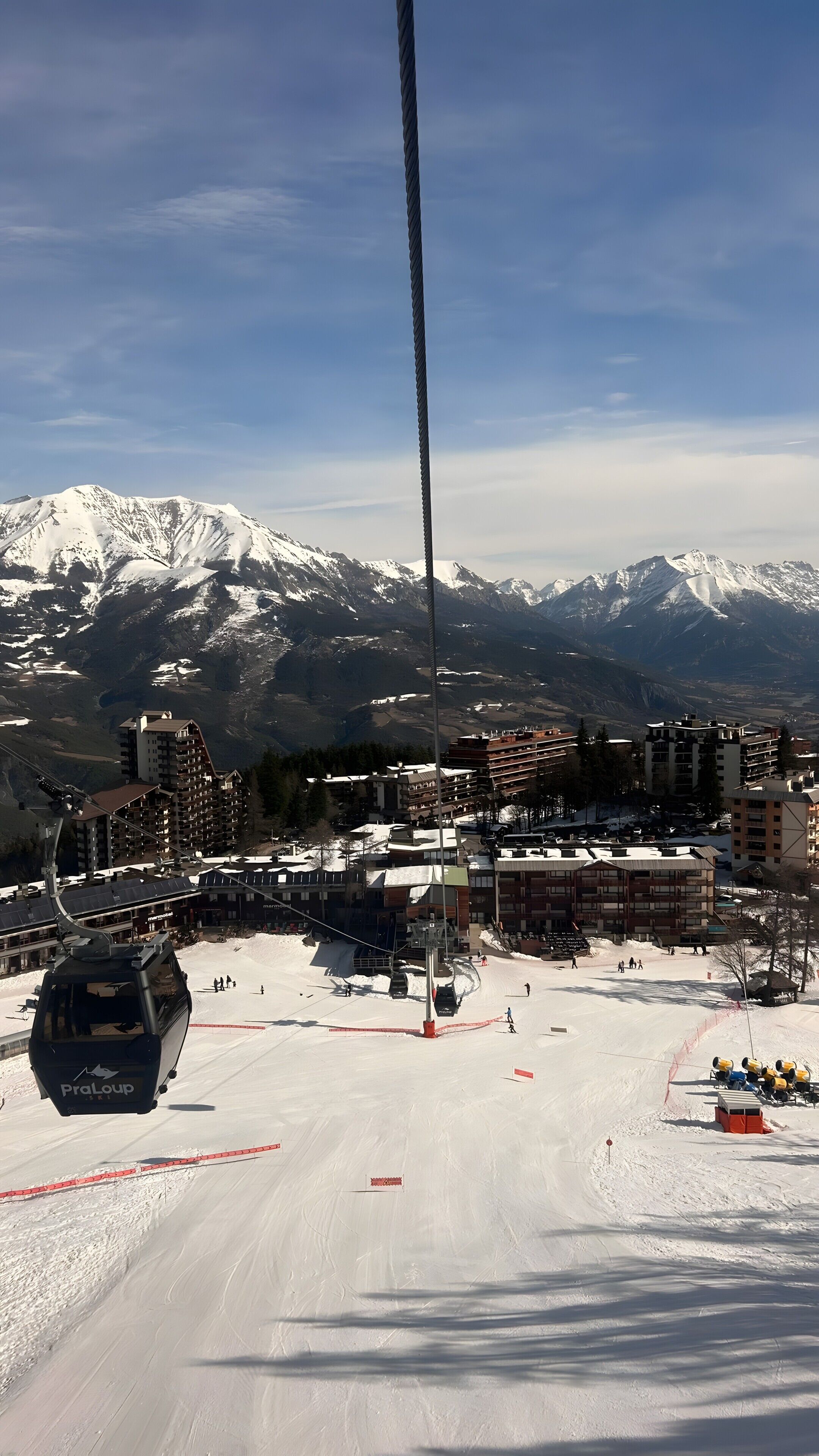 A ski lift with a cable, a snow-covered mountain range, and buildings in the background.