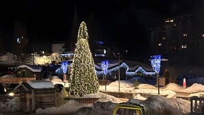 Ein verschneiter Außenbereich mit einem großen Weihnachtsbaum, einem gelben Fahrzeug und beleuchteten Gebäuden im Hintergrund.