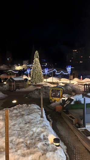 A snowy outdoor area with a large Christmas tree, a yellow vehicle, and illuminated buildings in the background.