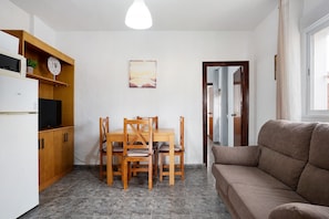A compact living space with a kitchenette, a dining table with chairs, a sofa, a painting on the wall, and a door leading to another room.