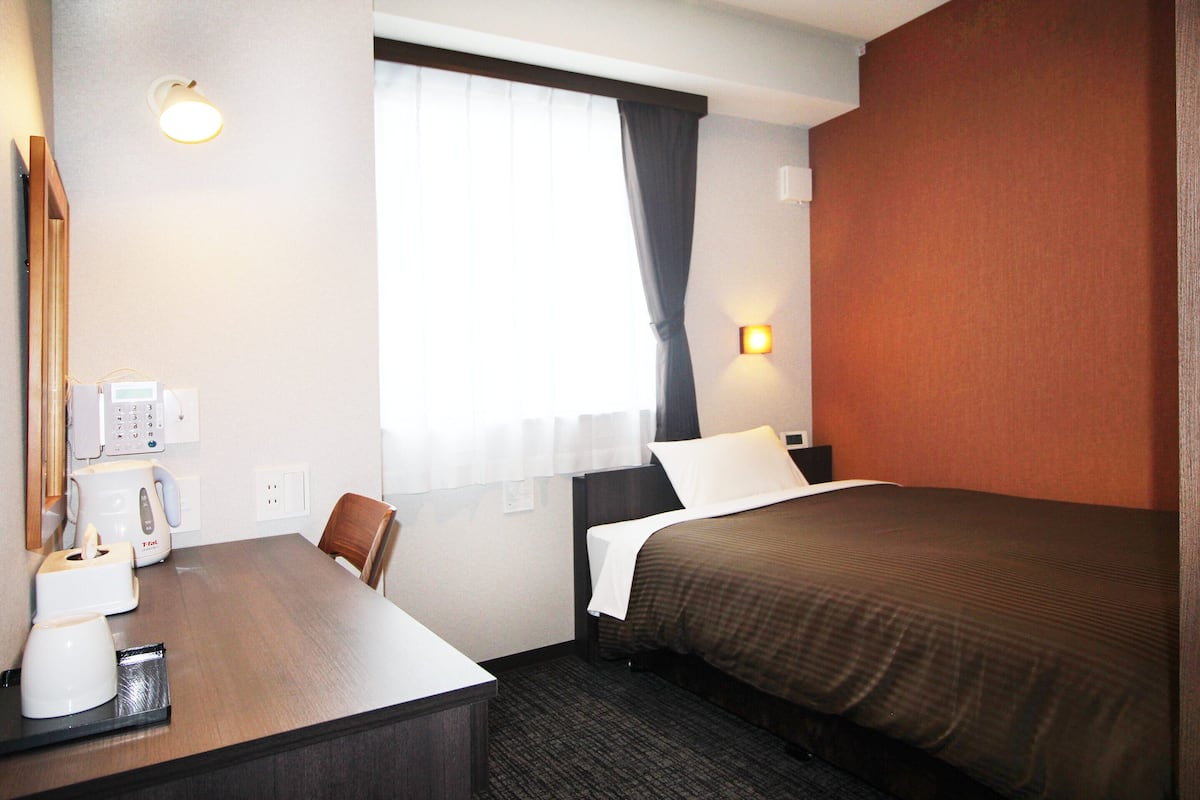 A hotel room with a bed, a desk with a coffee maker, a chair, a window with curtains, and a wall-mounted light.