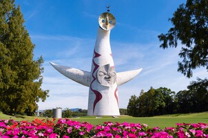 Una gran escultura abstracta con rostro, ubicada en un parque con césped verde y flores rosadas.