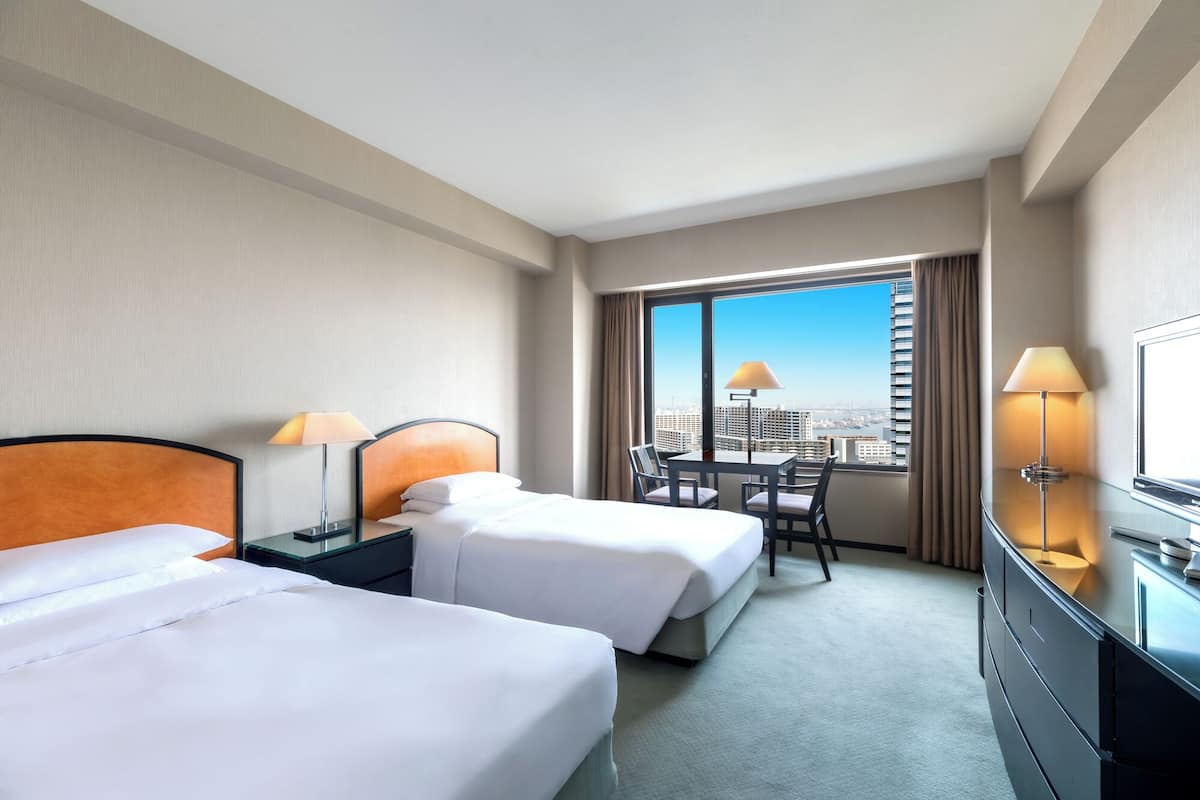 A hotel room with two beds, a desk, a chair, a TV, and a large window with a city view.