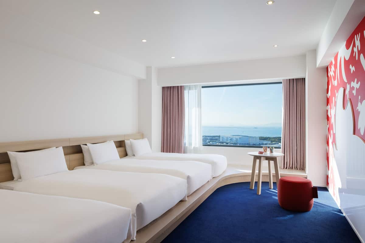A hotel room with three beds, a small table, a red ottoman, and a large window offering a city view.