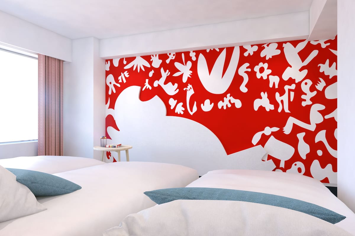 A hotel room with two beds, a red and white wall mural, a small table with a lamp, and a window with curtains.