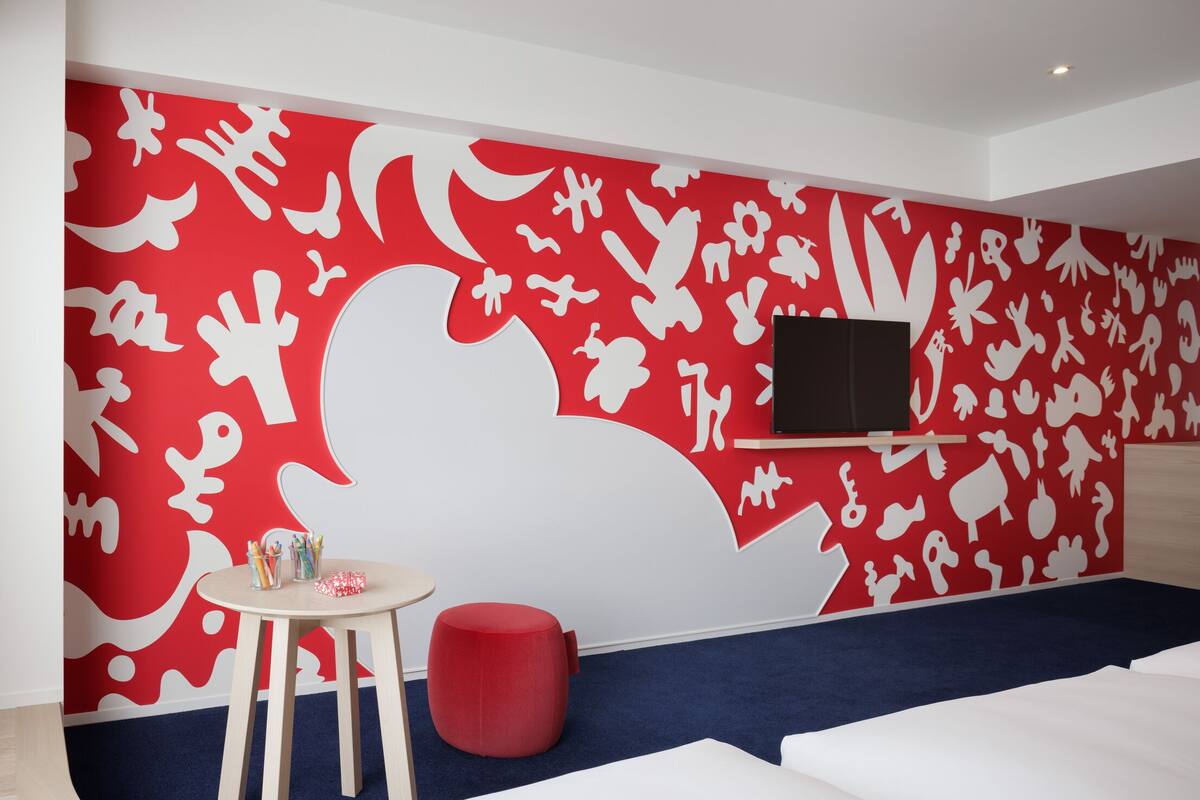 A room with a red and white abstract wall design, a flat-screen TV mounted on the wall, a small round red ottoman, and a white sofa.