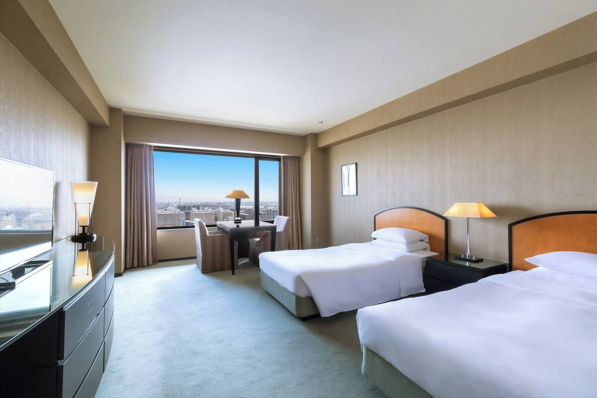 A hotel room with two beds, a desk, a TV, and a large window with a city view.