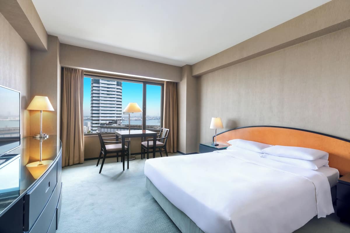 A hotel room with a bed, a desk with a chair, a TV, and a view of a cityscape.