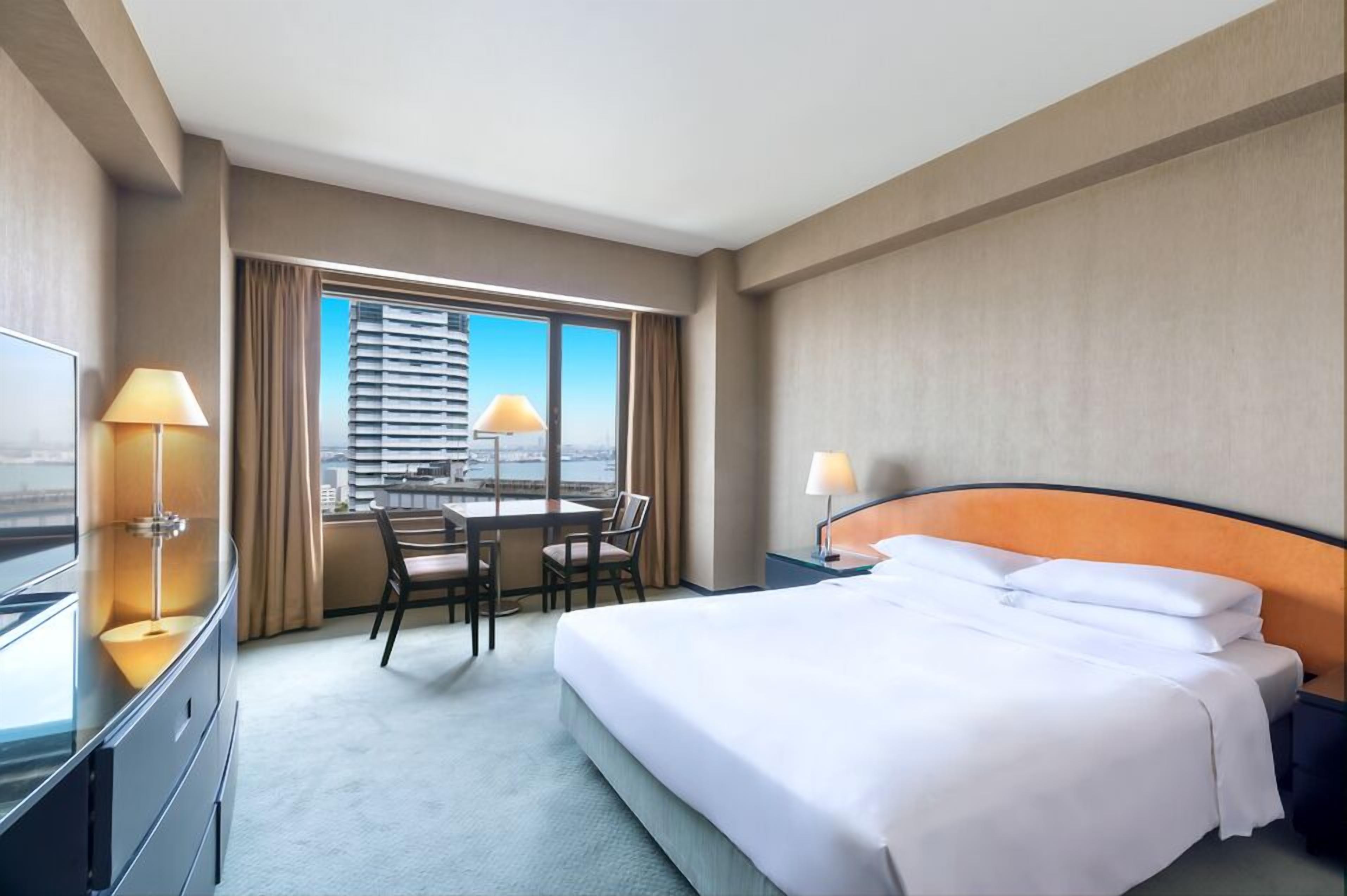 A hotel room with a bed, a desk with chairs, a TV, and a view of a cityscape.